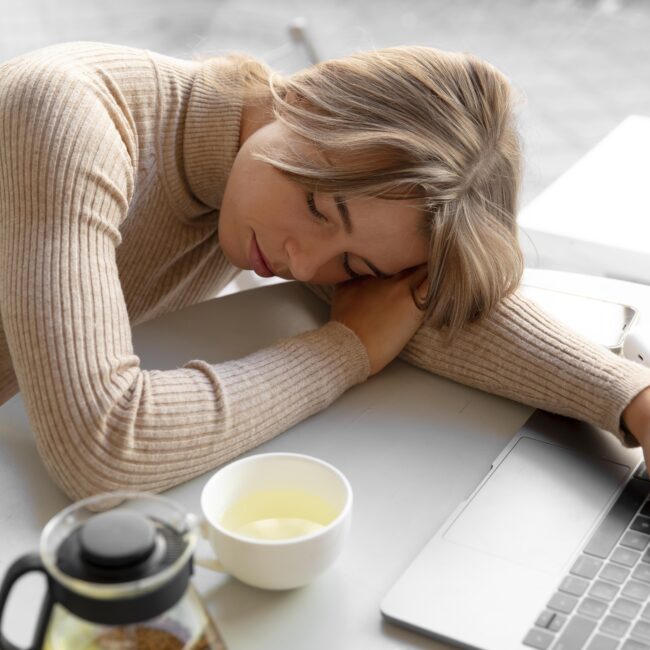 Alt : Une jeune femme est allongée sur un canapé dans une position détendue, les yeux fermés, son visage exprimant un soulagement après avoir été exposée à une surcharge de bruits. Ses mains sont positionnées délicatement près de ses oreilles, comme pour symboliser le besoin de silence et de repos auditif. La lumière douce et tamisée de la pièce renforce l'ambiance apaisante. Un casque anti-bruit repose sur une table basse à proximité, suggérant qu'elle vient de prendre une pause nécessaire face à la fatigue auditive. Son expression paisible contraste avec la tension précédente causée par le surmenage sonore.