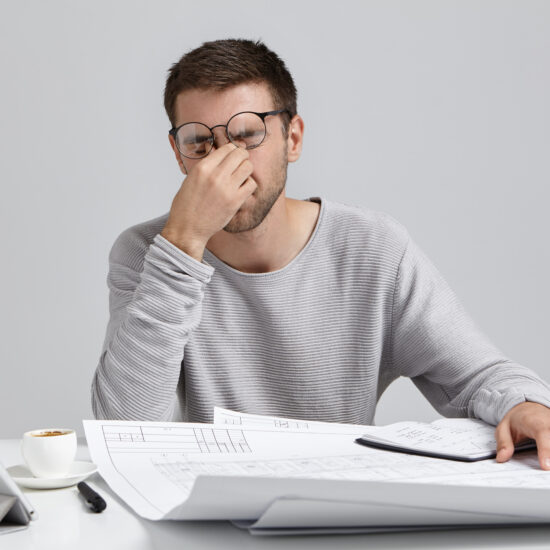 Stress, surmenage et délais. Jeune architecte indépendant fatigué et mal rasé massant l'arête du nez, ayant des maux de tête alors qu'il travaillait sur un grand projet architectural au bureau à domicile, se sentant épuisé et somnolent