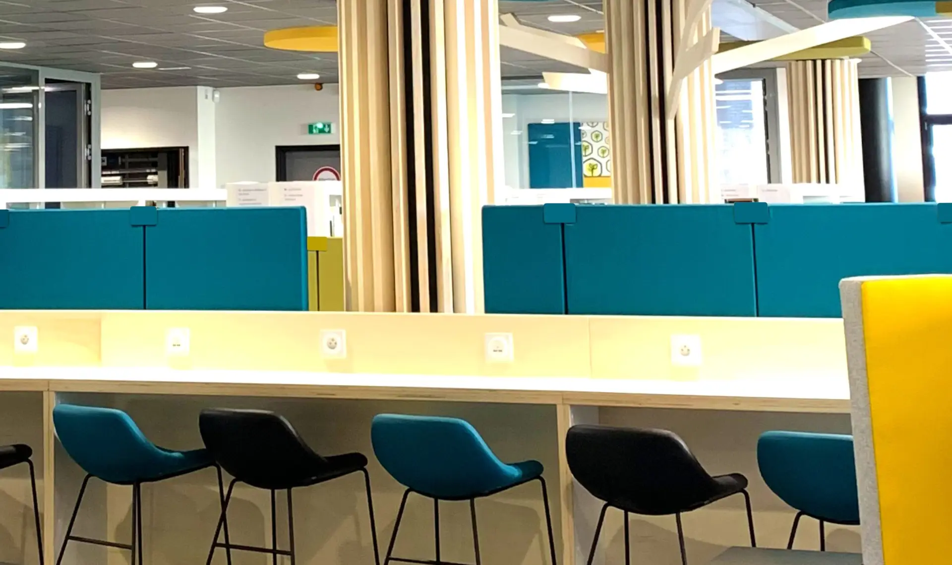 Open space moderne avec plusieurs bureaux partagés, équipé de panneaux acoustiques suspendus au plafond et de cloisons phonoabsorbantes colorées entre les postes de travail pour réduire le bruit. Des employés travaillent dans un environnement lumineux et calme, amélioré par les solutions acoustiques.